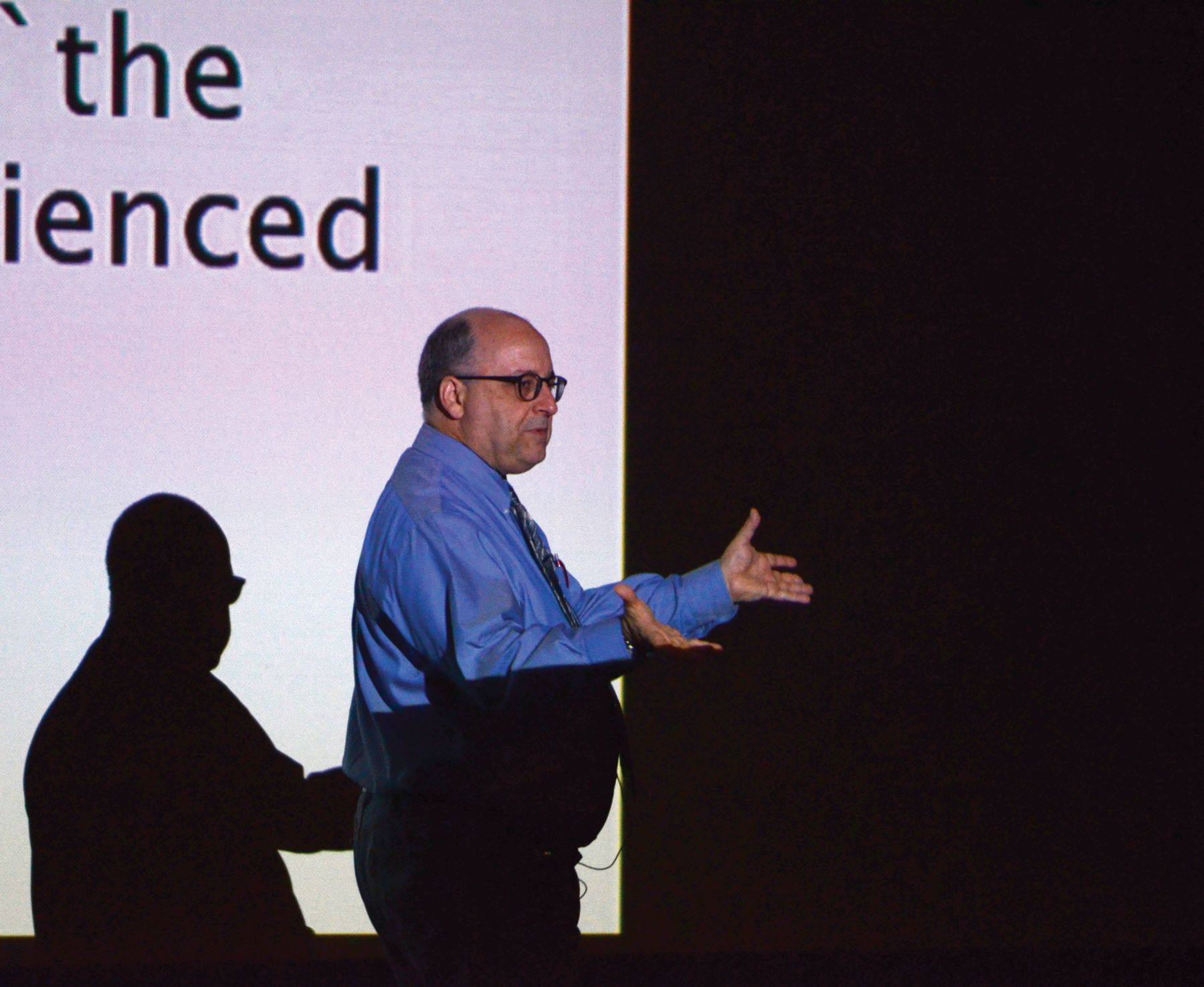 Faculty member Kevin Hagopian in front of a projector screen background gestures during a lecture