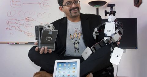 Portrait of S. Shyam Sundar, staged with several gadgets/pieces of technology while seated on desk.