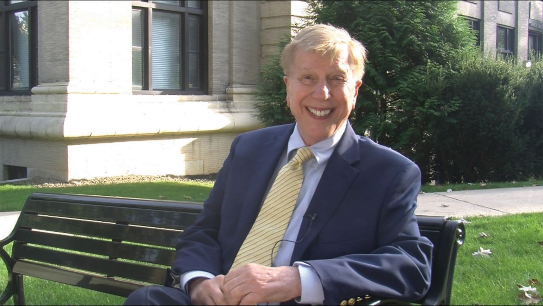 Jim Jimirro dressed in blue blazer with a yellow ties sits on a bench outside the Carnegie Building