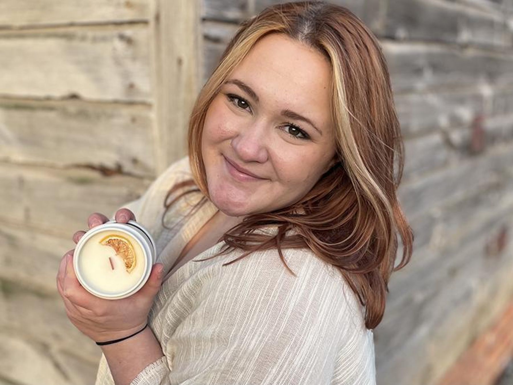 A smiling girl with wavy brown hair looks at the camera over her left shoulder while holding a candle in a glass jar.