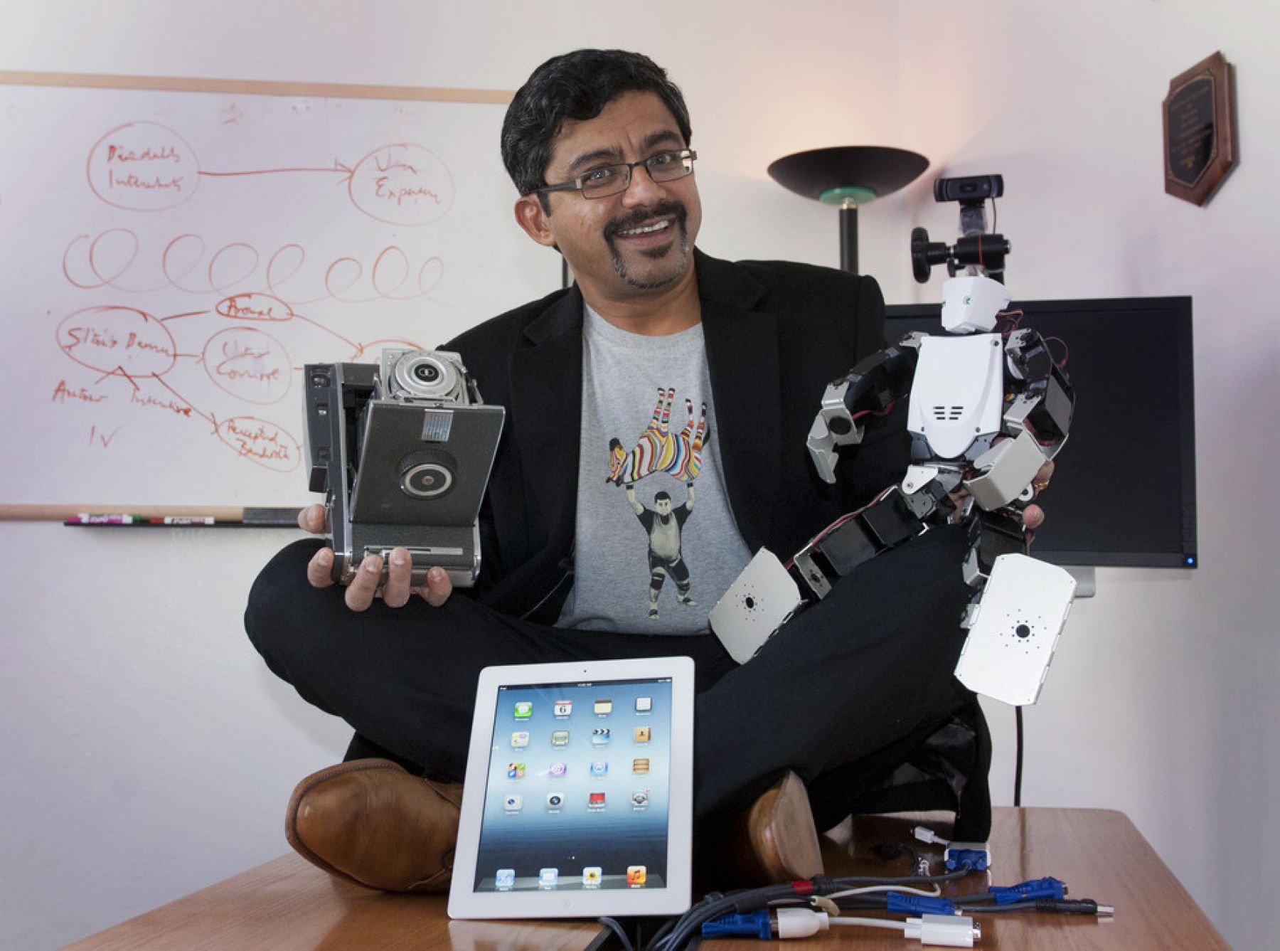 Portrait of S. Shyam Sundar, staged with several gadgets/pieces of technology while seated on desk.