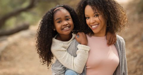 Dawn Manning poses in a pink shirt and gray sweater with her daughter hanging off her right shoulder