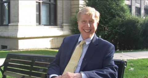 Jim Jimirro dressed in blue blazer with a yellow ties sits on a bench outside the Carnegie Building