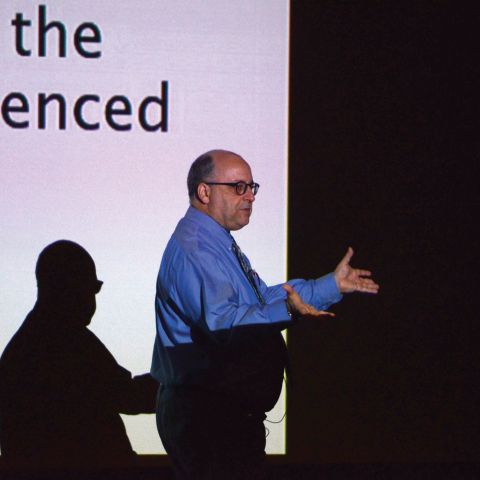 Faculty member Kevin Hagopian in front of a projector screen background gestures during a lecture